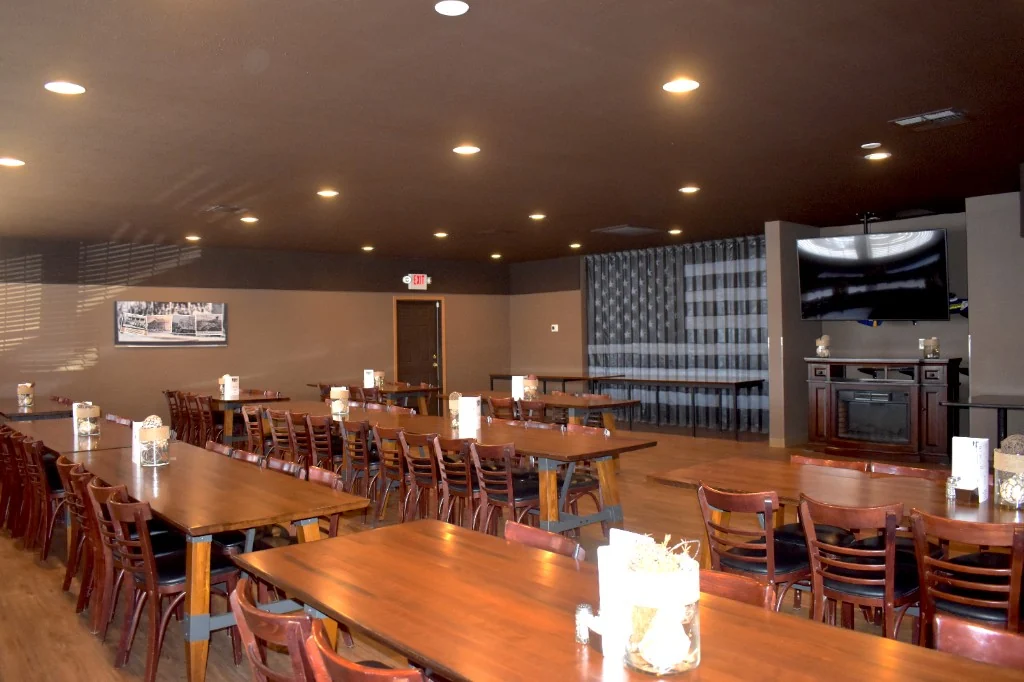 The banquet room with long wooden tables, warm recessed lighting, and an American flag on the back wall